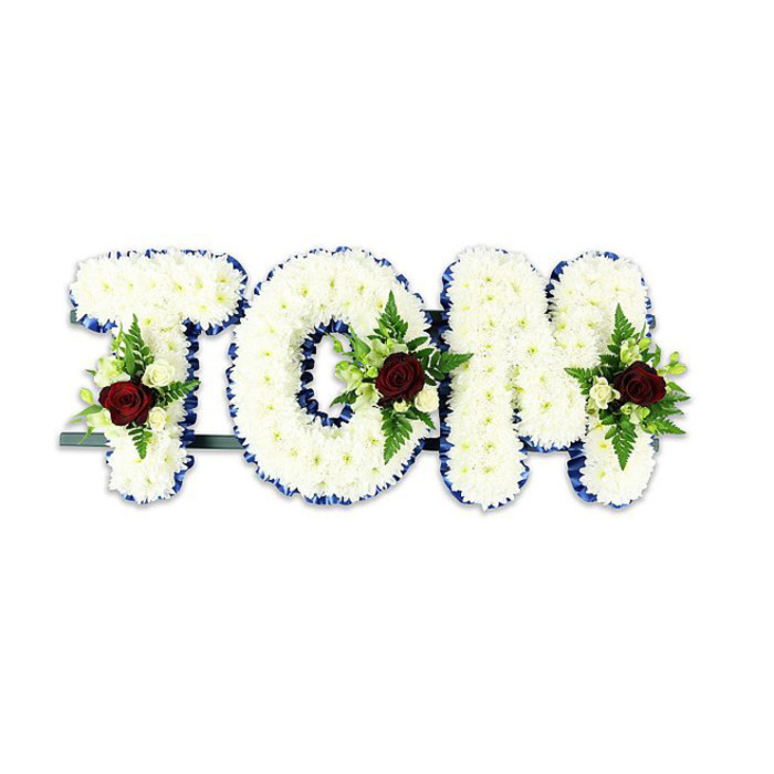 A formal, personalised memorial arrangement presents the name 'TOM' in bold, sculpted letters, each layer of the letters densely covered with tiny white chrysanthemum-like blooms to form a soft, cloud-like surface. The chrysanthemums' many petals create a tactile, layered texture that reads as both pure and comforting; in neutral light their subtle contours and shadows give each letter graceful depth. The edges of every letter are neatly finished with a vivid royal blue ribbon, the colour offering a crisp, elegant border which enhances legibility and lends a composed dignity. Placed thoughtfully into each letter, small floral sprays act as sombre accents: a single dark red rose anchors each spray, symbolising profound love and respect, surrounded by several smaller creamy white spray roses and sprigs of fern and leafy greenery for contrast and natural structure. A thin dark metal frame runs behind and beneath the letters, slightly visible at the base, indicating the piece is designed for upright display at a chapel, crematorium or funeral venue. Set against a plain white backdrop, the arrangement's whites, blues, reds and greens read cleanly and respectfully. This type of bespoke name tribute is commonly chosen for memorial services across West Ham and neighbouring communities - near Barking Road, Canning Town and local churches - where families look for handcrafted floral expressions that combine visual clarity with heartfelt sentiment. The composition is calm, meticulously balanced, and intended to offer solace through considered floral craftsmanship.