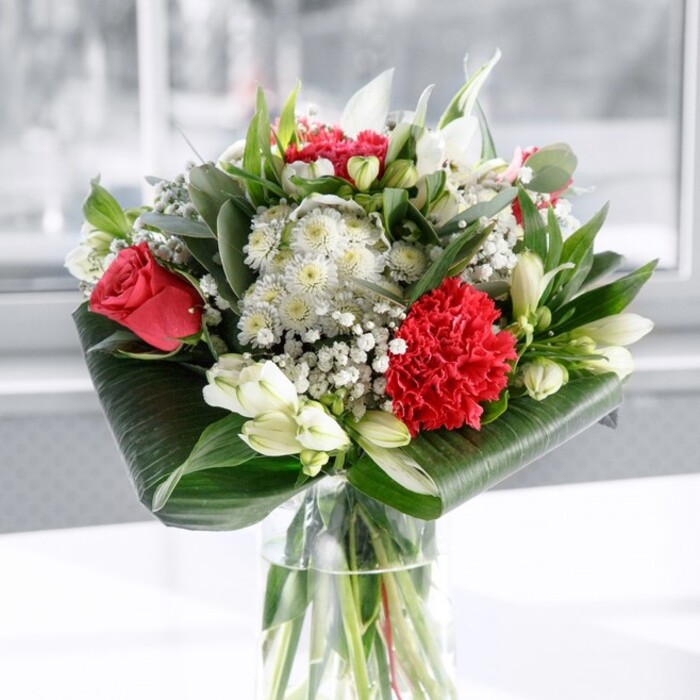 In this image, a carefully designed red and white bouquet stands in a clear cylindrical glass vase, the stems crisply cut and aligned, visible beneath the surface of clean water like the strong foundation of a relationship being celebrated somewhere in West Ham. At the centre, deep ruby roses with velvety petals open in soft spirals, surrounded by a luxuriously frilled crimson carnation whose densely layered petals catch the light and add drama. Around these focal blooms, creamy white alstroemeria with subtle speckles open gently, joined by white chrysanthemums and a scattering of lily buds, their green-tipped, tightly closed forms hinting at blossoms still to come. Sprays of gypsophila, or baby's breath, weave between the larger flowers, forming a delicate halo of tiny white dots that gives the arrangement a cloud-like softness. The greenery plays an equal part in the artistry: broad, glossy dark green leaves are wrapped and curved into a neat collar around the base of the bouquet, while narrower, pointed leaves and eucalyptus-style sprigs rise through the centre, adding height, contrast, and a sense of garden freshness. All of this rests on a white surface against a light, softly blurred background, evoking a bright East London morning-perhaps the kind of daylight that falls through the windows of a townhouse near West Ham Park or a cosy flat off Barking Road. The bouquet feels both contemporary and timeless, ideal as a dining table centrepiece, reception display, or bedside surprise. Its harmonious mix of bold red tones and calm whites makes it perfect for anniversaries, birthdays, or moments when you want to express deep gratitude or enduring love with flowers that look freshly arranged by a local expert florist.