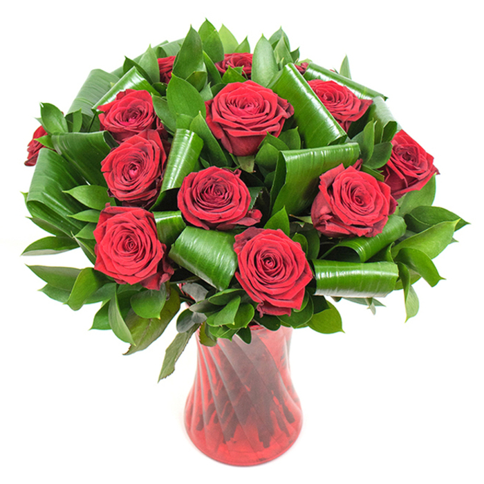 A carefully arranged cluster of deep crimson roses occupies the center of the photograph, each bloom fully open and revealing velvety, spiralled petals that create a dense, tactile surface of layered red. The roses number roughly a dozen or slightly more and are closely packed to form a lush, dome-like silhouette that reads as both generous and composed. Surrounding the rose heads is abundant dark green foliage: compact, glossy leaves form a rich backdrop while larger, bright green leaves - likely aspidistra-style - have been skillfully folded, rolled and spiraled into elegant cylinders and arcs. Those sculptural green elements lend a contemporary architectural rhythm to the classic rose bouquet, cradling blossoms and adding measured negative space so each head remains distinct. The stems rest in a tall, translucent ruby-red glass vase whose subtle twisted or ribbed texture creates vertical highlights that catch the soft, even lighting used here; the vase's slender, flared profile complements the rounded mass of flowers and anchors the design with a refined silhouette. Set against a pristine white studio background, the arrangement's colors and textures pop with clarity: the deep reds feel warm and saturated, the greens cool and verdant, and the glass offers a glossy, reflective counterpoint. The image conveys measured romance and craftsmanship - the kind of bouquet you might choose as an anniversary centrepiece or a considered Valentine's gift delivered to a flat near Green Street market or a townhouse close to Upton Park, offering both visual drama and the comforting scent of fresh roses.