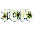 A personalised sympathy tribute in the image spells out the name "TOM" in three large floral letters, arranged side by side on a slim black frame set against a clear white background. Every letter is made up of tightly packed white chrysanthemums, their small rounded blooms overlapping so completely that the eye reads a smooth, plush surface of pure white. Running neatly along the outside edge of each letter is a trim of royal blue satin ribbon, precisely folded and pinned to follow every angle and curve, creating a striking border that cleanly defines the shape of the T, the rounded O, and the angular M. At the top left of the T, on the curve of the O, and at the upper right of the M, small but vivid sprays of flowers add depth and colour: deep red roses, velvety and full, sit amongst clusters of tiny cream or white spray roses and slender white flowers reminiscent of freesia. These are framed by sprigs of fern and other mixed greenery, whose fresh green tones help soften the strong white background and bring a touch of natural movement to the display. The tribute is carefully designed to be read clearly during a funeral or memorial service in and around West Ham, perhaps resting near the front of a chapel or beside the hearse. It is both traditional and deeply personal, with the name Tom spelt out in flowers that speak of love, remembrance, and the gentle support offered to family and friends in their time of loss.